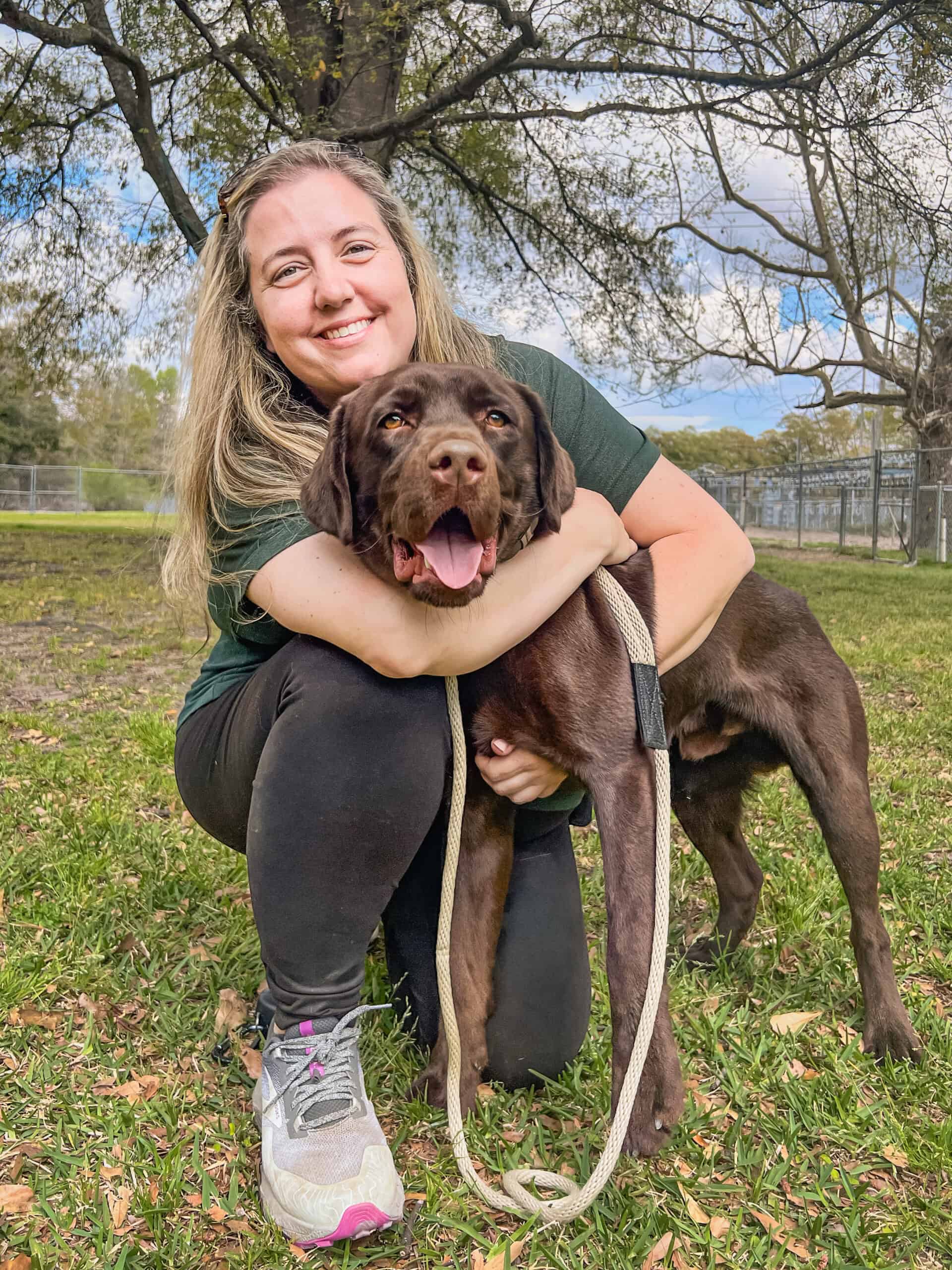 Janie Wilkerson, co-owner of Well Mannered Dog, kneeling and hugging her chocolate Labrador Hopper outdoors