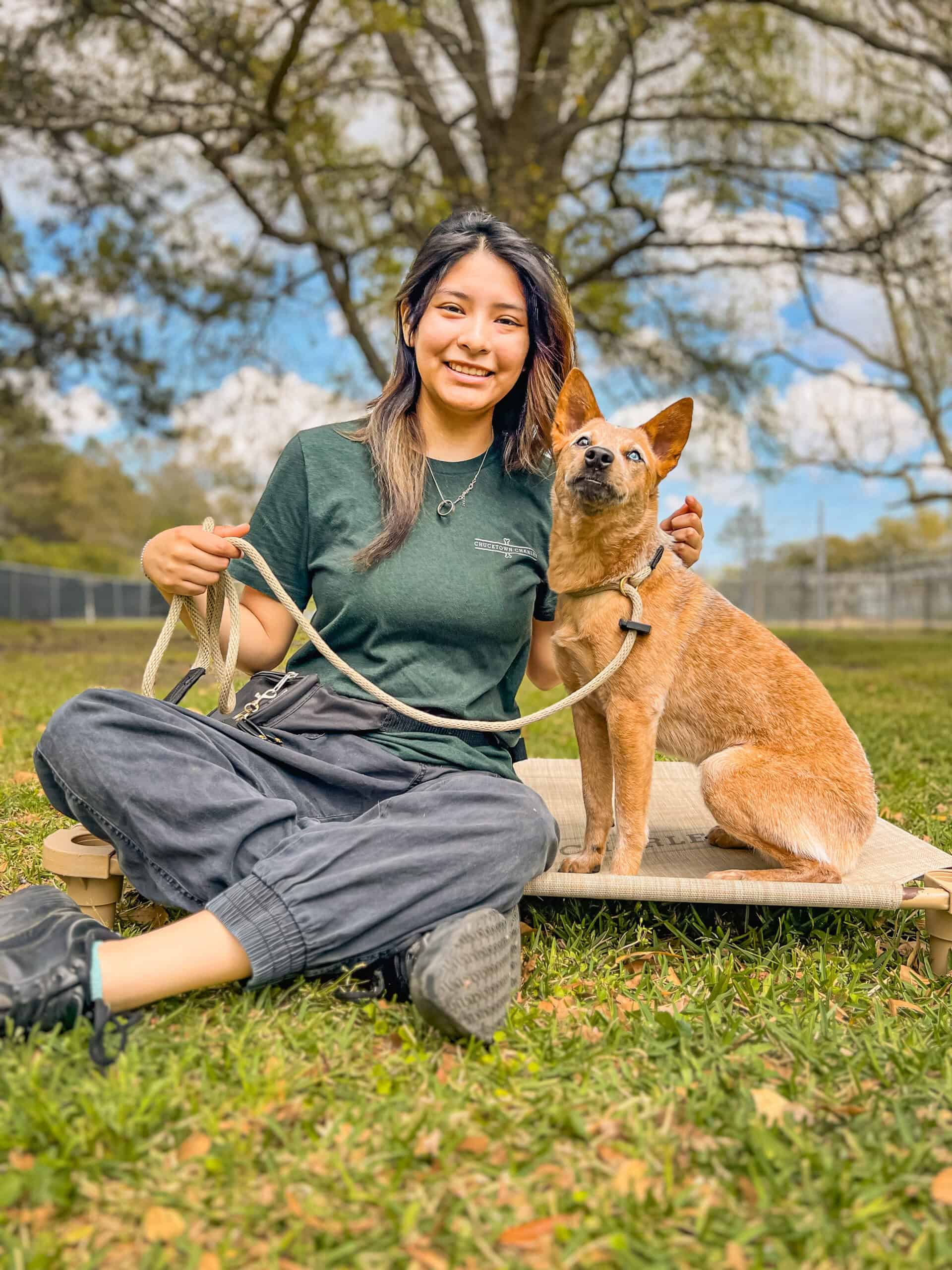 Lesly Salgado of Well Mannered Dog sitting outdoors beside a red heeler on a raised cot